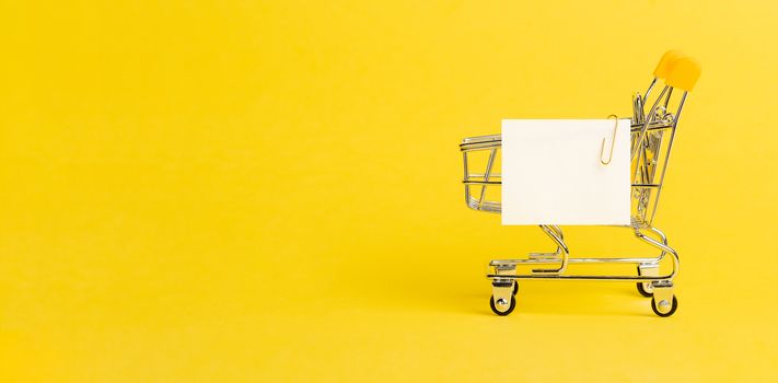 Shopping cart and white paper note list over yellow background. Shopping concept on bright yellow background. Empty white paper note over shopping cart. Copy space for text or design. Banner