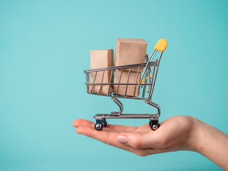 Toy shopping cart with boxes in female hand over blue background. Copy space for text or design. Ssale, discount, shopaholism concept. Consumer society trend
