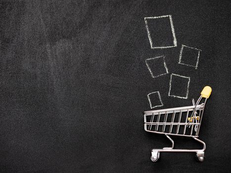 Shopping cart and products on chalkboard. Miniature supermarket shop trolley as shopaholism concept with copy space left. Top view or flat lay.
