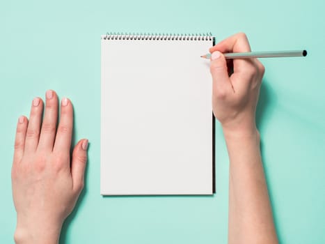Blank paper notebook and female hands with pencil. Empty paper sketchbook with pincil in woman hands on blue background. Top view or flat lay. Copy space for text or design.