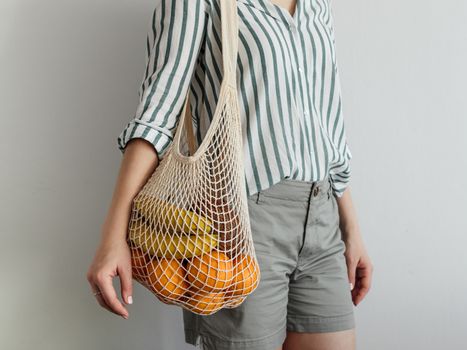 Mesh bag with fruits on female shoulder. Stylish young woman standing with mesh shopping bag on shoulder near light wall. Modern reusable shopping concept.