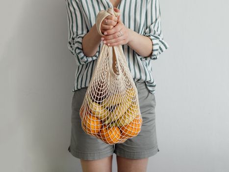 Mesh bag with fruits in female hands. Stylish young woman hold mesh shopping bag on light gray wall. Modern reusable shopping concept.
