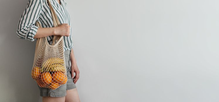 Mesh bag with fruits on female shoulder. Stylish young woman standing with mesh shopping bag on shoulder near light wall. Modern reusable shopping concept, copy space.