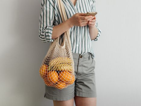 Unrecognizable young woman using smartphone standing with mesh bag on shoulder. Stylish young woman standing with mesh shopping bag on shoulder near light wall and using smartphone.