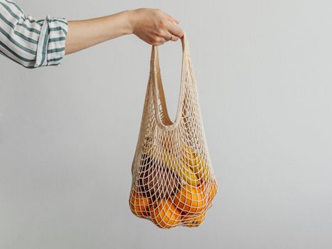 Mesh bag with fruits in female hand. Hand of Stylish young woman hold mesh shopping bag on light gray wall. Modern reusable shopping concept.