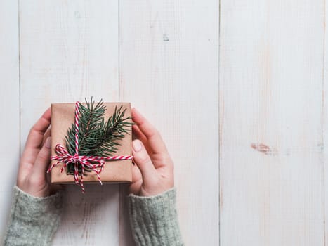 Female hands hold gift box over white wooden background, copy space right. Caucasian girl hands in gray sweater holding gift box in craft wrapping paper with red Christmas string and fir branches