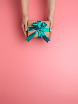 Female hands hold gift box on pink background, copy space down. Caucasian girl hands holding gift box in craft wrapping paper with green satin ribbon. Christmas, New Year or Birthday present