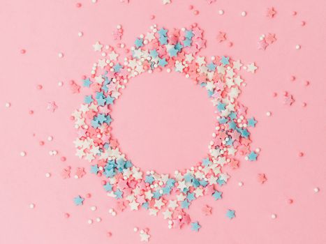 Festive border frame of colorful pastel sprinkles on pink background with copy space in center. Sugar sprinkle dots and stars in round shape, decoration for cake and bakery. Top view or flat lay