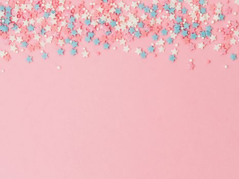 Festive border frame of colorful pastel sprinkles on pink background, copy space top. Sugar sprinkle dots and stars, decoration for cake and bakery. Top view or flat lay