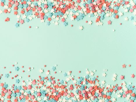 Festive border frame of colorful pastel sprinkles on blue background with copy space in center. Sugar sprinkle dots and stars, decoration for cake and bakery. Top view or flat lay.