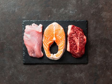 Raw turkey breast fillet, salmon steak and beef ribeye steak on black slate board over dark background. Top view or flat lay. Protein food, keto, carnivore, paleo diet concept.