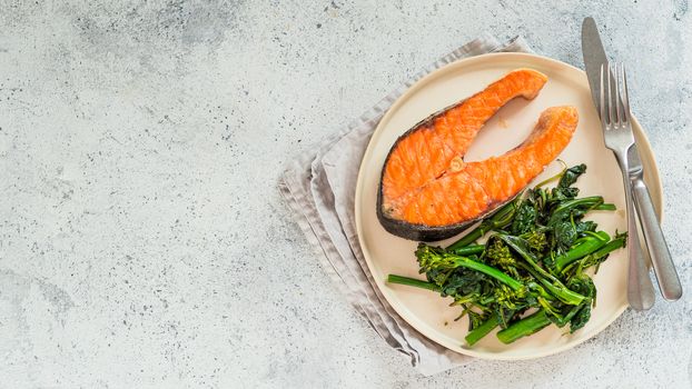Ready-to-eat grilled salmon steak and greens - baby broccoli or broccolini and spinach on rustic craft plate over gray background. Keto diet dish. Top view or flat lay.Copy space left for text. Banner