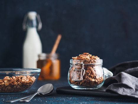 Homemade granola in glass jar on dark table. Ingredients for healthy breakfast - granola, milk and honey. Copy space for text. Low key.