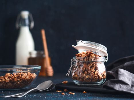 Homemade granola in glass jar on dark table. Ingredients for healthy breakfast - granola, milk and honey. Copy space for text. Low key.