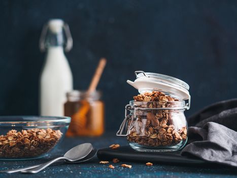 Homemade granola in glass jar on dark table. Ingredients for healthy breakfast - granola, milk and honey. Copy space for text. Low key.