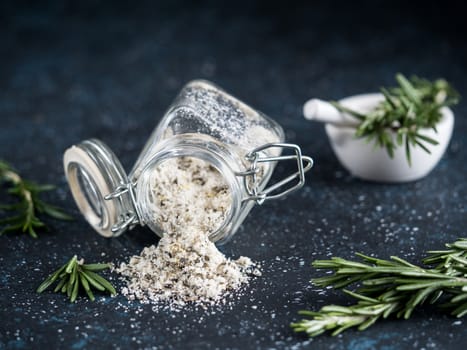 Sea salt scented herb rosemary and lemon zest. Sea salt with aromatic herb spilled out of glass jar on dark blue background. Scented salt and ingerdients. Copy space
