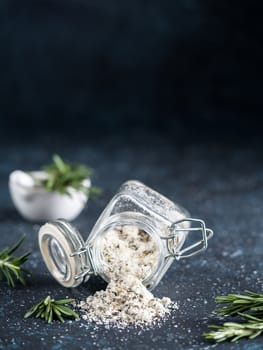 Sea salt scented herb rosemary and lemon zest. Sea salt with aromatic herb spilled out of glass jar on dark blue background. Scented salt and ingerdients. Copy space