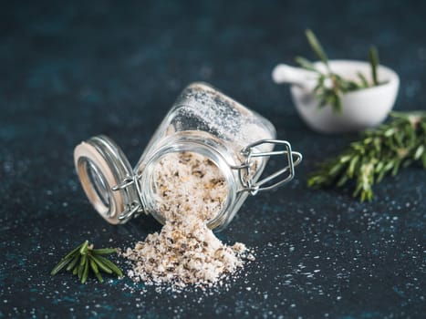 Sea salt scented herb rosemary and lemon zest. Sea salt with aromatic herb spilled out of glass jar on dark blue background. Scented salt and ingerdients. Copy space