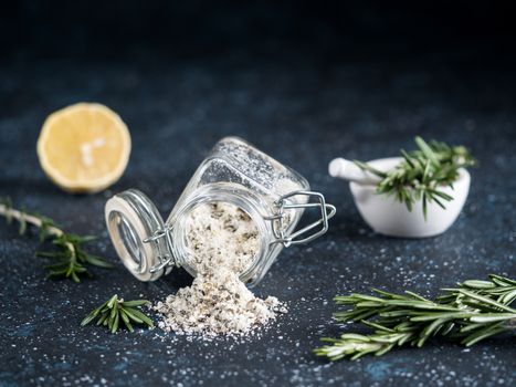 Sea salt scented herb rosemary and lemon zest. Sea salt with aromatic herb spilled out of glass jar on dark blue background. Scented salt and ingerdients - fresh rosemary and lemon. Copy space