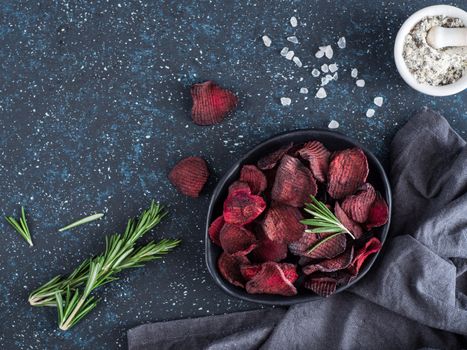 Baked beet slices with coarse sea salt scented rosemary. Vegan diet food idea and recipe. Healthy homemade beetroot chips in plate and ingredients on dark blue background. Copy space. Top view