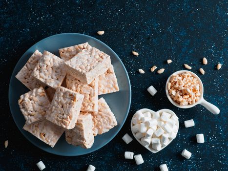 Homemade square bars of Marshmallow and crispy rice and ingredients on dark blue background. American dessert with marshmallow and crispy rice. Top view. Copy space