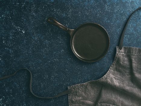 Cooking and kitchen background - trendy denim cotton apron and empty iron-cast pan on dark table. Gray kitchen apron on dark table as kitchen background with copy space for text. Top view or flat-lay