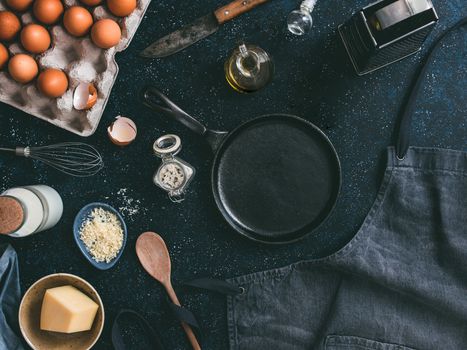 Empty cast-iron frying pan and ingredients for omlette or scrambled eggs,top view.Egg,shell,milk,parmesan cheese,scented rosemary salt,olive oil,pepper,apron,whisk,vintage knife ondark table.Copyspace