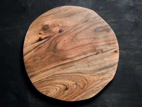Round wooden tray or cutting board on black table. Top view of empty kitchen trendy rustic wooden tray saw cut imitation on black wooden background. Copy space for text. Food and menu background.
