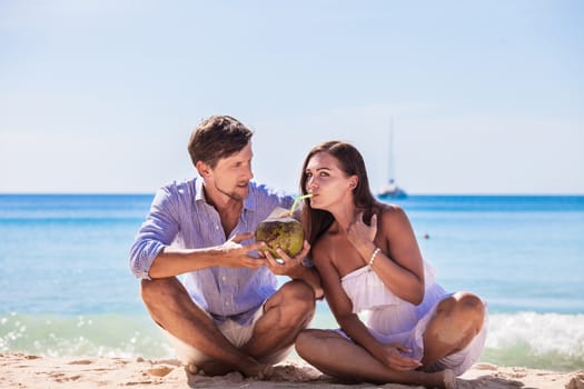 Cheerful couple sitting on beach and drinking coconut milk