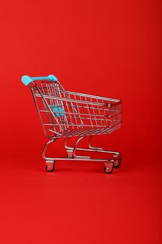 Close up empty toy metal supermarket shopping cart over vivid red background with copy space, low angle side view