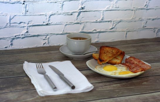 A plate with two fried eggs, slices of bacon and two toasts is on the table, next to a cup of black coffee and cutlery.