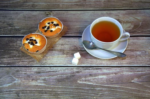 A cup of tea with two sugar cubes and a spoon on a saucer are on the table next to two chocolate muffins on a textile napkin.