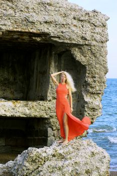 Tanned pretty blonde girl in a red dress stands on the stone ruins of an old fort, against the backdrop of the waves of the Baltic Sea.