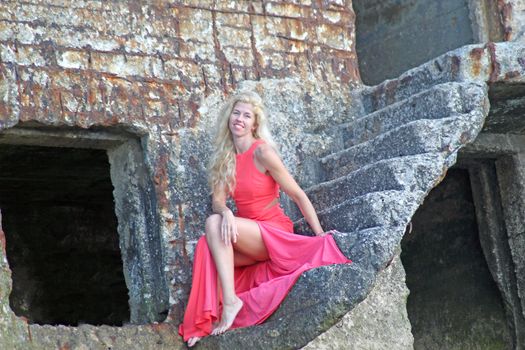 Tanned pretty blonde girl in a red dress sitting on the stone ruins of an old fort.