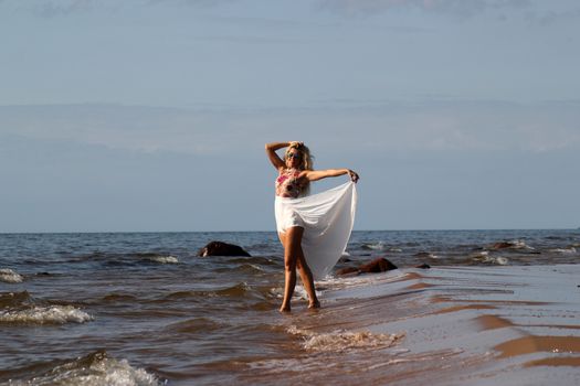 tanned blonde in white dress walks on the water on the beach of the Baltic sea on a sunny day.