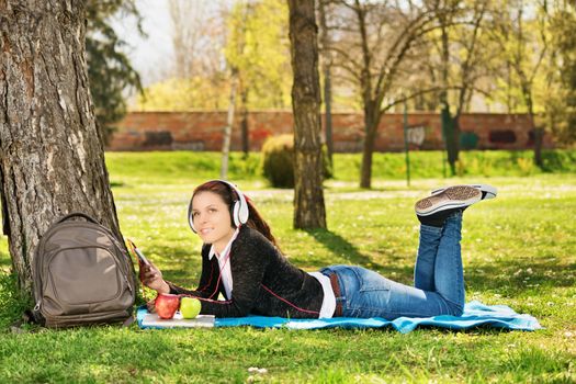 A portrait of a beautiful female student, lying on a blanket on the grass in a park listening to music. Student taking a break from studying.