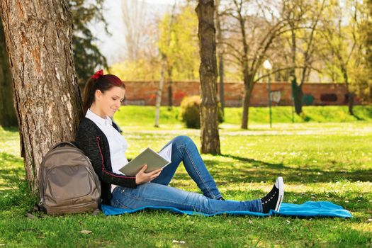 A portrait of a beautiful female student in a park, sitting on the grass, reading a book. Girl studying for her exams in the park.