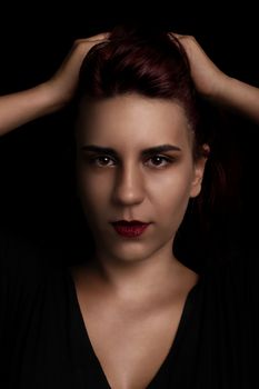 Low key sensual beauty portrait of a red haired young woman holding her hair on black background.