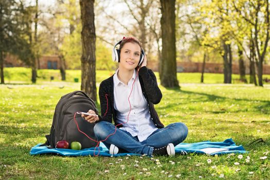 Beautiful young female student sitting on the grass in a park, listening to music, making a break from her studying.