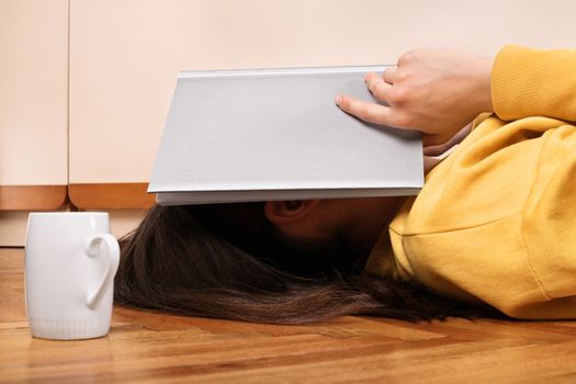 Late night study. Young girl lying on the floor in her room with a book over her head next to a coffee cup. Bored of reading. 