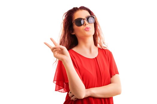 Young beautiful redhead girl in a red blouse with sunglasses gesturing the peace sign and sending a kiss, isolated on a white background.