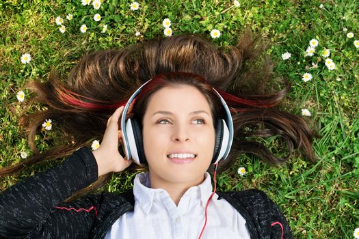 A close up portrait of a beautiful young girl lying on the grass in a park, listening to music on her headphones. 