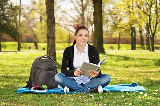 A portrait of a beautiful young female student holding a book, sitting on the grass in a park.