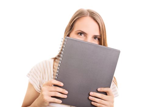 Beautiful girl hiding her smile behind a notebook, isolated on white background. Girl with a mischievous look hiding behind her notebook.