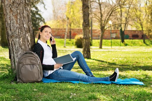 Beautiful young female student sitting in a park, leaning on a tree, talking on the phone. Young girl making a break from her studying.
