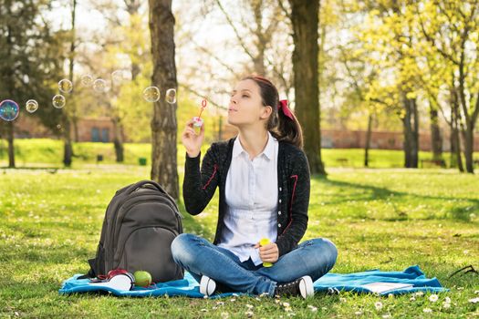 Dream of wishes coming through. Beautiful young student girl sitting on a meadow blowing soap bubbles, taking a break from studying.