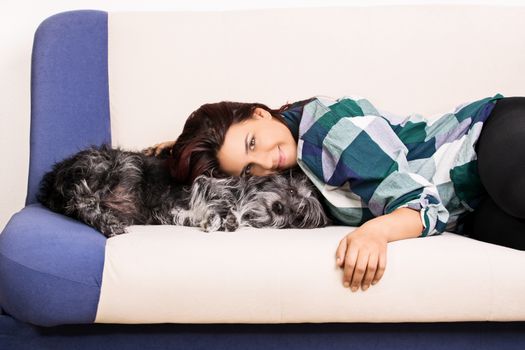 Beautiful young smiling girl, lying on a couch at home and hugging her cute little dog.
