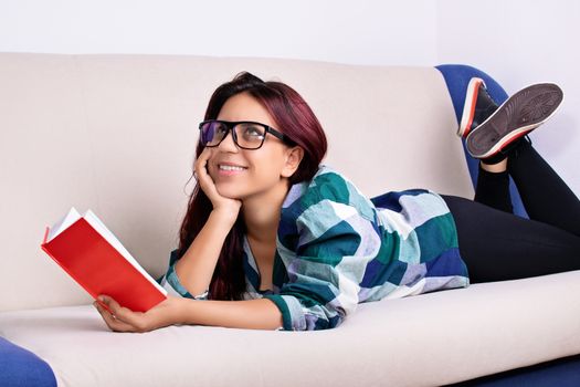 A close-up shot of a young bookaholic girl with glasses reading a book at home, looking up and daydreaming.