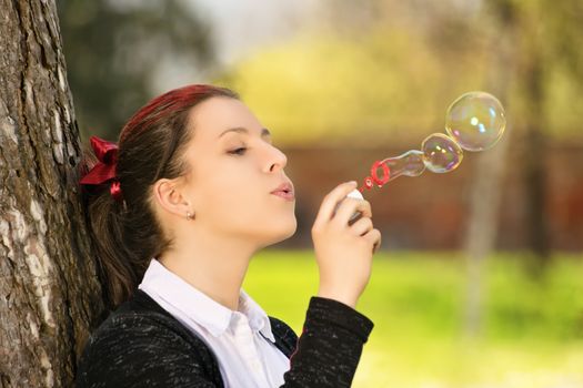 A close up portrait shot of a beautiful young student girl in a park, leaning against a tree, blowing soap bubbles.