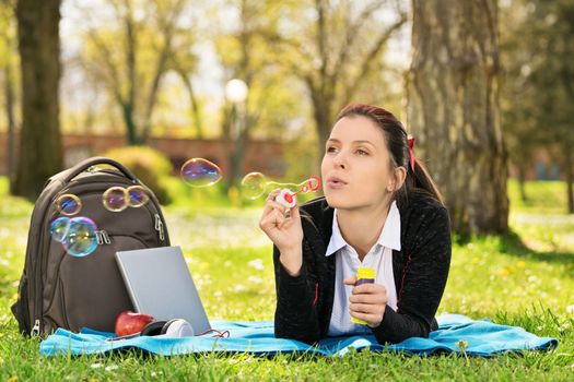 Blowing soap bubbles in the wind. Close up shot of a beautiful young student girl lying on a meadow blowing soap bubbles, taking a break from studying.